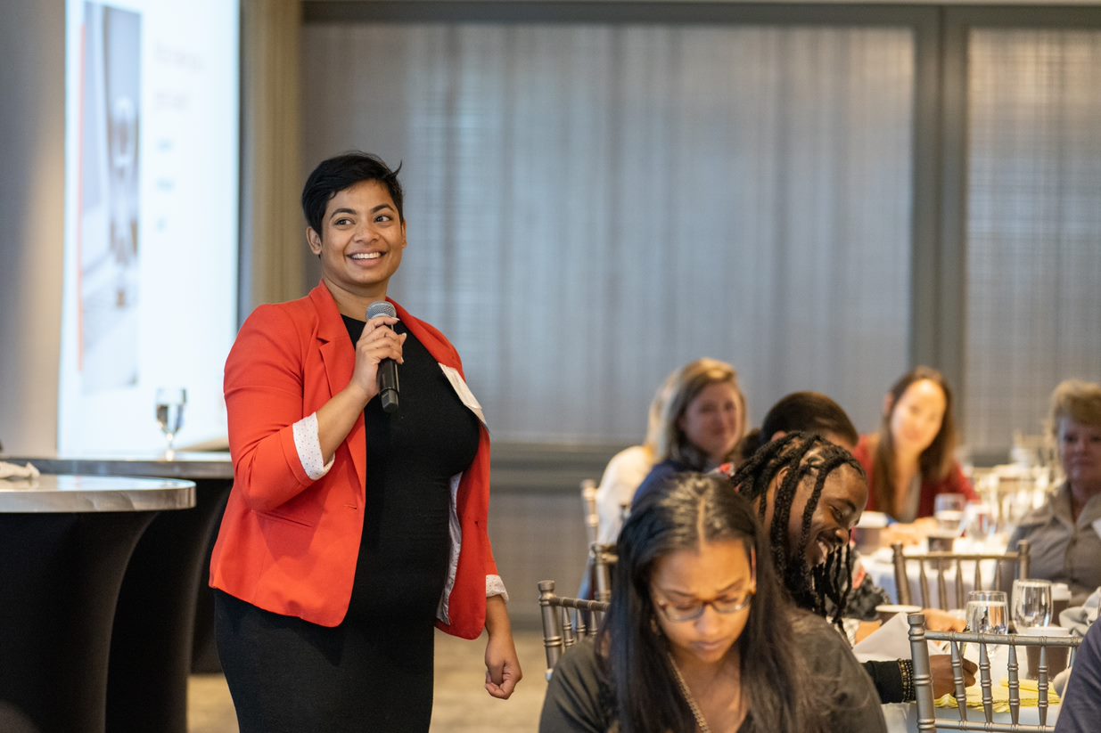 A woman in a orange jacket talking on a microphone to a group of people