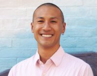 An Asian man with close-cut hair and a light pink button-down shirt smiles in front of a blue brick wall.