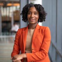 A Black woman in a orange blazer stands in a hallway with a relaxed smile on her face.