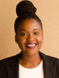 A Black woman with braids put up in a bun smiles. She is wearing a black blazer with a white t-shirt underneath.